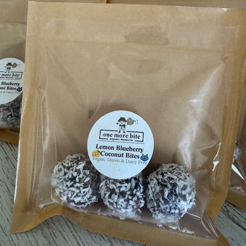 Package of three gluten-free, Dairy-Free and Vegan Lemon Blueberry Coconut Bites made with organic almonds and pecans, sweetened with dates. Three coconut bites displayed with a scattering of dates, almonds, pecans and dried blueberries.