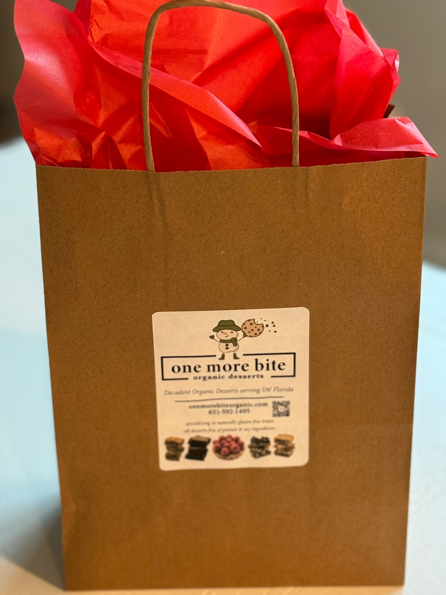 A sample of a kraft handle gift bag with white One More Bite Organic label and red tissue paper