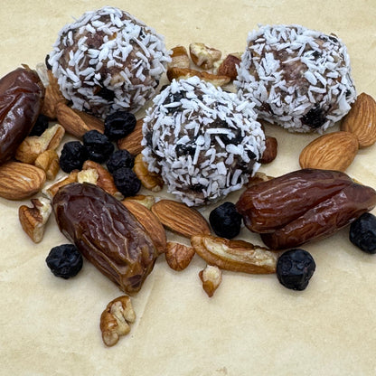 Gluten-free, Dairy-Free and Vegan Lemon Blueberry Coconut Bites made with organic almonds and pecans, sweetened with dates. Three coconut bites displayed with a scattering of dates, almonds, pecans and dried blueberries. 