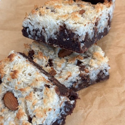 Gluten-free, organic Almond Joy Brownie made with a fudgy brownie base, topped with a coconut macaroon layer and an organic almond shown as a stack of three decadent brownie treats.